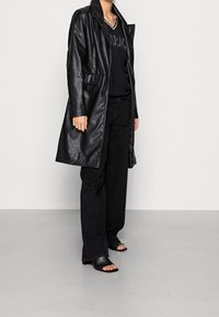 Osoba ubrana w długi czarny skórzany płaszcz, czarne spodnie o szerokich nogawkach, czarną bluzkę z dekoltem w serek z logo oraz czarne buty na obcasie z odkrytymi palcami, stojąca na szarej podłodze.