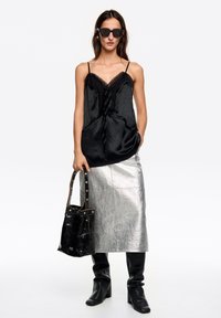 Czarna satynowa kamizelka z koronkowym wykończeniem, zestawiona ze srebrną metaliczną spódnicą midi, czarną torebką i czarnymi kozakami za kolano.