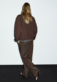 Giacca bomber marrone trapuntata con spalle basse, indossata sopra un maglione grigio testurizzato e pantaloni larghi marroni, vista da dietro.