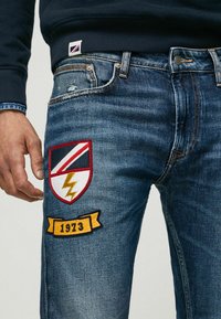 Jean en denim bleu avec une texture usée, présentant une broderie rouge et jaune représentant un blason et l'année "1973".