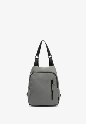 Mochila gris con un exterior liso y resistente al agua. Cuenta con correas ajustables negras, un bolsillo frontal con cremallera y una forma redondeada.