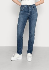 Jeans azuis de cintura alta com corte justo, leve desgaste e ligeira agressão. Combinados com ténis brancos para contraste.