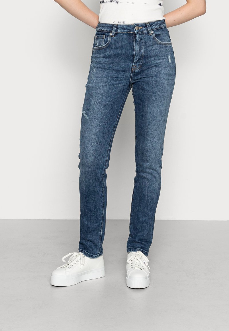 Jeans azuis de cintura alta com corte justo, leve desgaste e ligeira agressão. Combinados com ténis brancos para contraste.