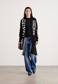 Črn cardigan z belim logotipom "DKNY", v kombinaciji s črno majico in širokimi modrimi kavbojkami. Značilni so ostro konicami črne čevlje in majhna črna torbica.