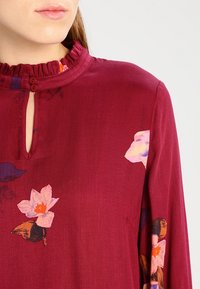 Bordeauxfarbende Bluse mit Blumenmuster, die einen gerafften Ausschnitt, einen Tropfenausschnitt und Knopfdetails aufweist. Lange Ärmel mit lebhaften Blumenakzenten.