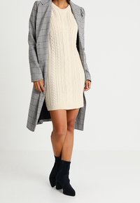 Beige pletené šaty se vzorem copánků, spárované se šedým károvaným kabátem. Černé kotníkové boty dotahují vzhled a zdůrazňují vrstvený outfit.