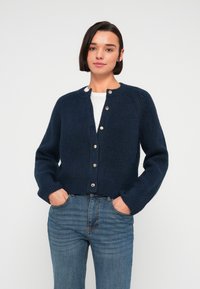 Μπλε σκούρο πλεκτό ζακέτα με στρογγυλή λαιμόκοψη, διακοσμημένη με χρυσά κουμπιά και ριμπ υφή. Συνδυάζεται με ανοιχτόχρωμα μπλε τζιν, με τα χέρια στις τσέπες.