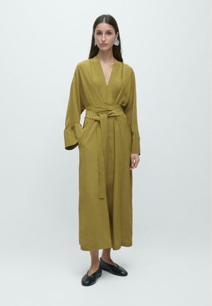 Kvinde iført olivengrøn lang kjole med bælte og brede ærmer samt sorte flade sko, stående mod en ensfarvet hvid baggrund.