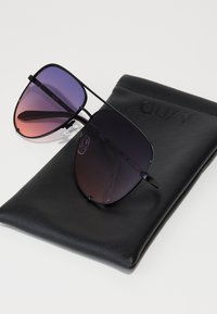 Schwarz gerahmte Sonnenbrille mit Verlaufsgläsern in lila und rosa Tönen, die auf einem schwarzen, weichen Etui mit dem eingeprägten Schriftzug "QUAY" liegen.