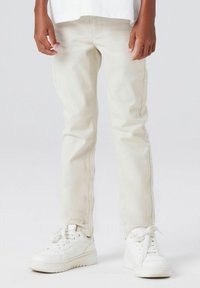 Beige denimbukser med smal passform, med en glatt tekstur og subtile sømmer. Kombinert med hvite joggesko, vist mot en grå bakgrunn.