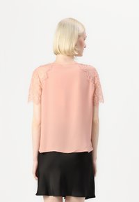 Ροζ μπλούζα με δαντελένια κοντά μανίκια και απαλή, ρέουσα σιλουέτα; η μαύρη φούστα από κάτω προσθέτει αντίθεση.