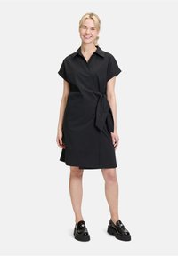 Schwarzes Wickelkleid mit kurzen Ärmeln, einem kragenlosen Ausschnitt und einer gebundenen Taille. Getragen mit glänzenden schwarzen Plateauschuhen und flachen Absätzen.
