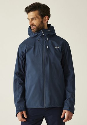 Homme portant une veste imperméable à capuche bleu marine avec une fermeture éclair frontale et une poche poitrine, regardant vers sa gauche.