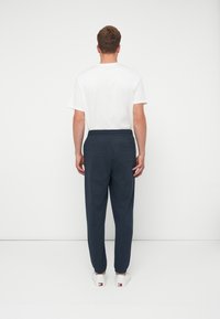 Bărbat îmbrăcat cu un tricou alb, pantaloni sport de culoare bleu închis și adidași albi, stând cu fața spre un zid alb simplu, având mâinile relaxate pe lângă corp.
