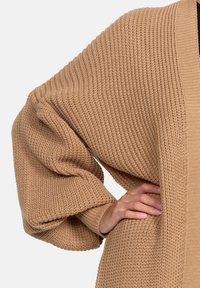 Beige överdimensionerad stickad cardigan med en texturerad vaffelmönster. Har vida ärmar och krage i sjalkonstruktion. Detalj av hand på tröjan.