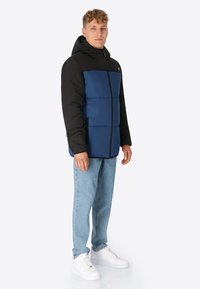 Gestepptes Jackett mit schwarzer Kapuze und Ärmeln, blauem Vorderteil, Reißverschluss und Seitentaschen. Getragen mit hellblauen Jeans und weißen Sneakers.