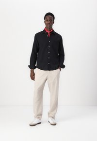 Camicia nera con bottoni e tasca, pantaloni di lino beige e un fazzoletto rosso attorno al collo. Sneaker bianche con suola marrone.
