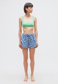 Γυναίκα με πράσινο αθλητικό σουτιέν και μπλε σορτς με λουλούδια, όρθια ξυπόλητη και κοιτάζει προς τα αριστερά της μπροστά σε απλό λευκό φόντο.