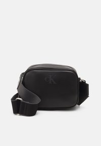 Černá kompaktní krosbody taška s nastavitelným širokým popruhem a embosovaným logem "cK" vpředu, hlavní přihrádka na zip.