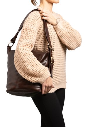 Braune Leder-Schultertasche mit glatter Textur, ausgestattet mit einem breiten Tragegurt und metallenen Hardware-Elementen. Tragen von einer Person in einem beigen Pullover.