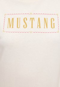 Light beige t-shirt featuring the word "MUSTANG" in bold gold letters, bordered with a decorative pink outline. Smooth cotton texture.