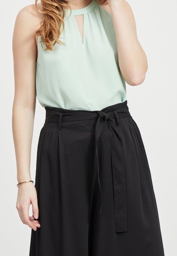 Femme portant un haut sans manches vert clair avec un décolleté en trou de serrure et un pantalon noir taille haute à jambes larges avec ceinture nouée.
