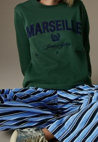 Person, die einen grünen Kapuzenpullover mit dem Schriftzug "MARSEILLE", gestreifte blaue und schwarze Hose und silberne Sneakers trägt, sitzt mit crossed legs.