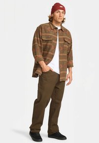 Camisa de botão castanha com padrão sobre uma t-shirt branca, combinada com calças castanhas e um gorro vermelho; sapatos pretos completam o visual.