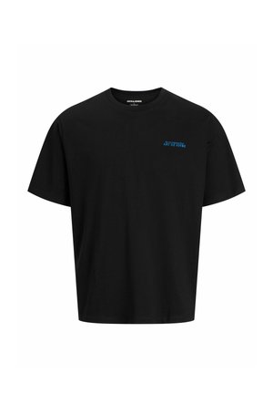 Ung person med krøllet hår iført solbriller, sort T-shirt, lyse denimshorts, hvide sokker og brune støvler, står foran en ensfarvet baggrund.
