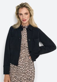 Veste noire à boutons avec col et deux poches avant, superposée à une robe à imprimé léopard. Texture lisse et coupe ajustée.