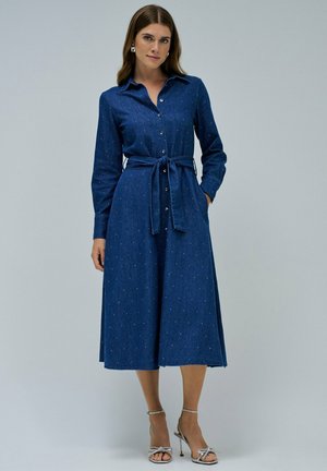 Femme portant une robe bleu marine à manches longues avec des petits pois blancs, une ceinture nouée et des sandales à talons aiguilles argentées, se tenant devant un fond uni.
