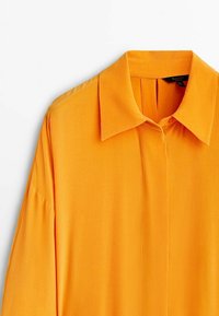 Langärmliges, mit Kragen versehenes Hemd aus leuchtend orangefarbenem Stoff. Verfügt über eine durchgehende Knopfleiste und Falten auf der Rückseite für zusätzliche Textur und Details.