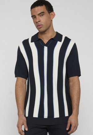 Mężczyzna ubrany w granatowy, krótki sweter z białymi i jasnoniebieskimi pionowymi paskami oraz kołnierzem na złożenie.