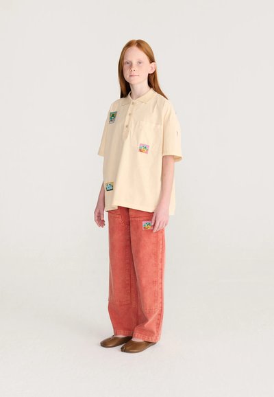 Jeune personne aux cheveux roux portant un polo crème oversize et un pantalon rouge ample avec des patchs colorés, debout sur un fond clair uni.