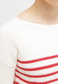 Weißer Strickpullover mit roten horizontalen Streifen. Gerippte Textur und runder Ausschnitt. Die Details umfassen einen kontrastierenden Abschluss an den Streifenrändern.