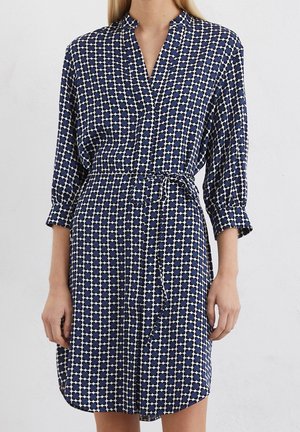 Femme portant une robe bleu et blanc à motifs, longueur genou, avec des manches trois-quarts et une ceinture nouée à la taille.