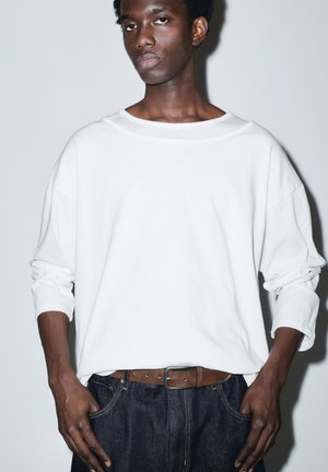 Fiatal férfi sötét bőrrel és rövid, göndör hajjal, laza fehér hosszú ujjú inget, barna övet és sötétkék farmert visel, egyszerű háttér előtt állva.