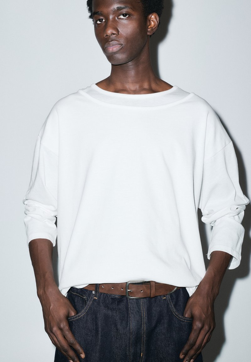 Giovane uomo con pelle scura e capelli corti e ricci, indossa una camicia bianca a maniche lunghe larga, cintura marrone e jeans blu scuro, in piedi contro uno sfondo semplice.