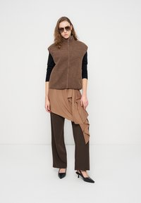 Καφέ γιλέκο από φλις με ψηλό γιακά, φορεμένο πάνω από τοπ σε χρώμα ταουπ με πτυχώσεις και καφέ παντελόνι με φαρδιά μπατζάκια. Μαύρες μυτερές γόβες και γυαλιά ηλίου ολοκληρώνουν την εμφάνιση.