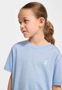 Ljust blå kortärmad t-shirt av mjukt tyg, med rundad hals och en liten vit grafik av tre chevroner på vänster bröst.