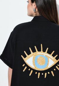 Czarna koszula z dużą złotą grafiką oka z niebieskimi akcentami i promieniującymi liniami na plecach. Teksturowana tkanina i luźny krój.