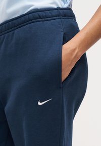 Άτομο που φοράει σκούρο μπλε φόρμα Nike με το χέρι στην τσέπη, δείχνοντας το λευκό κεντητό λογότυπο Nike swoosh κοντά στην τσέπη.