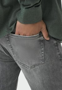Jeans in denim grigio con un design attillato, dotati di una tasca posteriore e un orlo leggermente sfilacciato. Indossati con una camicia verde foresta a bottoni.