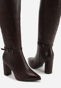 Bottes over-the-knee en cuir synthétique marron avec un bout pointu, un talon carré et une sangle à boucle ajustable à la cheville.