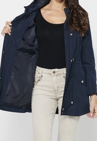 Femme portant une veste bleu marine avec fermeture éclair ouverte, un haut noir et un pantalon beige brodé, montrant la doublure intérieure sombre de la veste.
