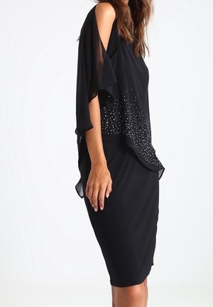 Woman wearing a black cold-shoulder dress with sheer fabric and scattered silver embellishments on the upper front.