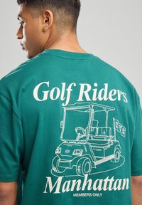 Vihreä puuvillapaita, jossa on valkoinen graafinen printti golfautosta ja teksti "Golf Riders Manhattan" selässä. Lyhyet hihat, pyöreä kaula-aukko.