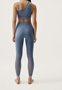 Kvinna står barfota, iförd blå höga leggings med genomskinliga nätpaneler och en matchande asymmetrisk sport-bh, sedd bakifrån.