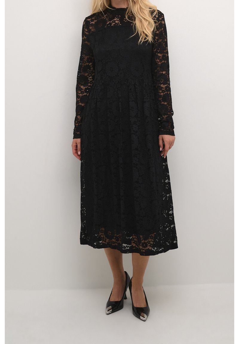 Femme portant une robe midi en dentelle noire à manches longues et des talons pointus noirs avec des embouts métalliques, debout sur un fond uni.