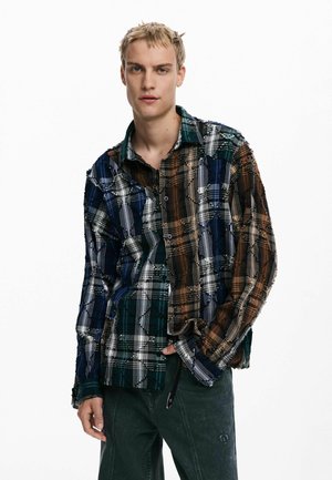 Checkered shirt in blue, green, brown, and white shades with a loose fit, frayed edges, and button closure. Worn over dark denim pants.